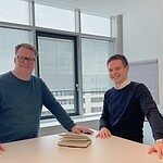 Zwei Männer im Büro bei der Arbeit, mit Notizbüchern auf einem Tisch.