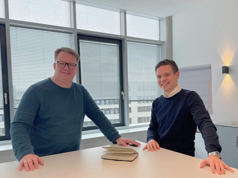 Zwei Männer im Büro bei der Arbeit, mit Notizbüchern auf einem Tisch.